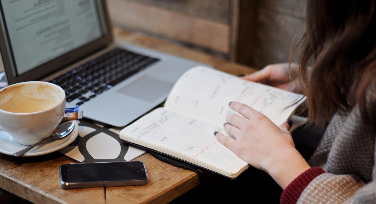 一个女人坐在咖啡馆里喝咖啡的特写照片, 一台笔记本电脑, 在她面前的桌子上有一个笔记本.