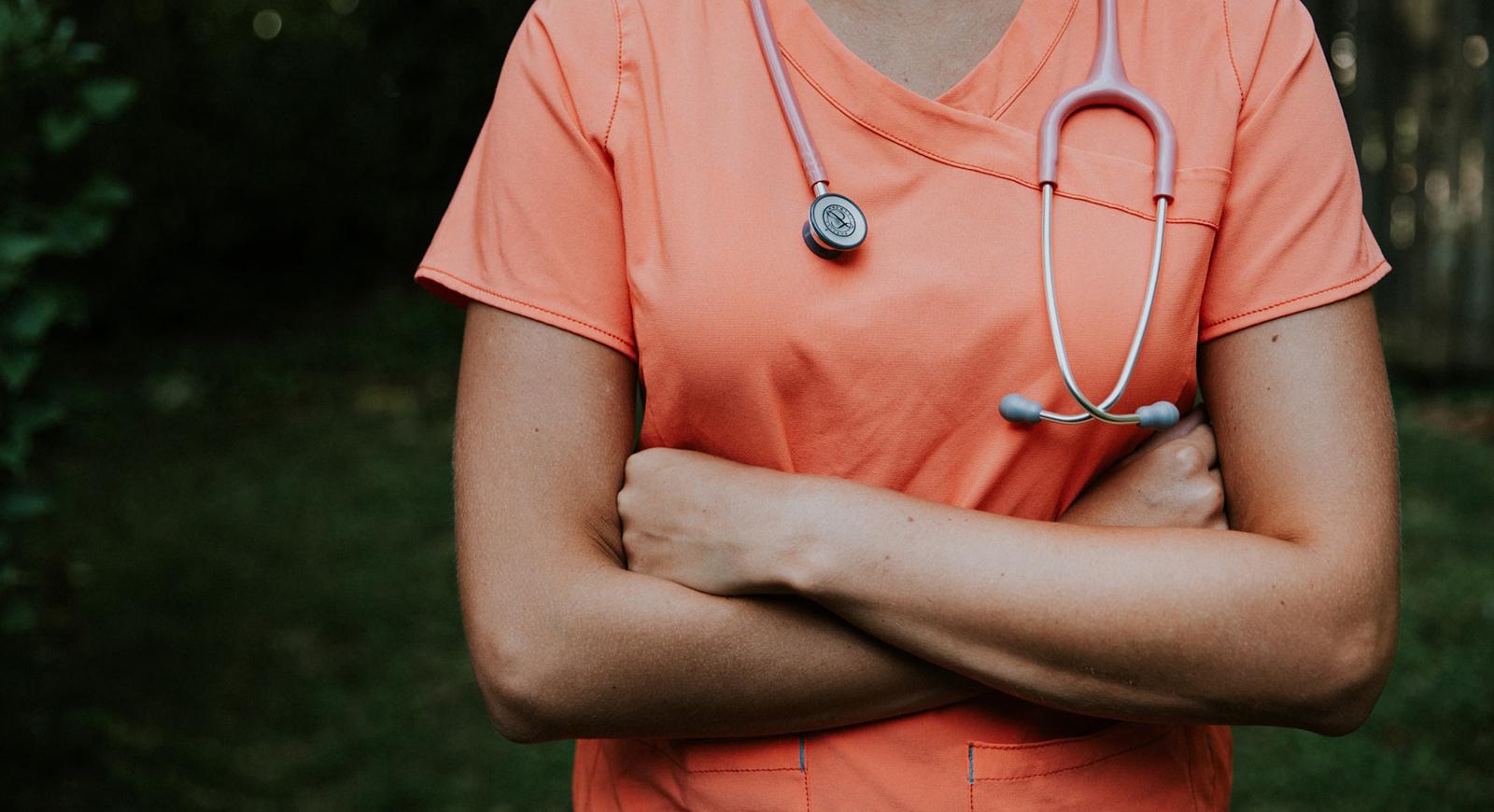 照片中，一名女护士穿着橙色工作服，戴着听诊器，双臂交叉站在绿色植物前.